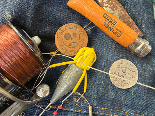 A fishing jig lure attached to a wire rig, displayed on a blue denim background alongside a spool of line, a wooden Opinel brand hook, and two circular metal pirate badges