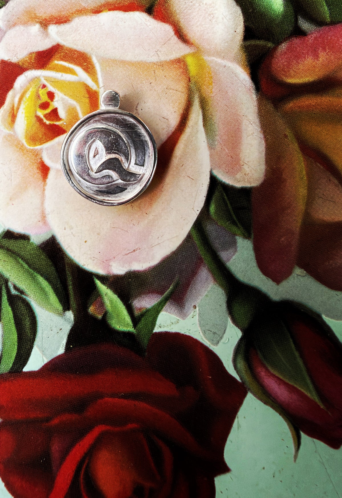A silver-toned circular pendant or charm with a dark emblem in the center, resting on a painted floral surface with cream, red, and green flowers.