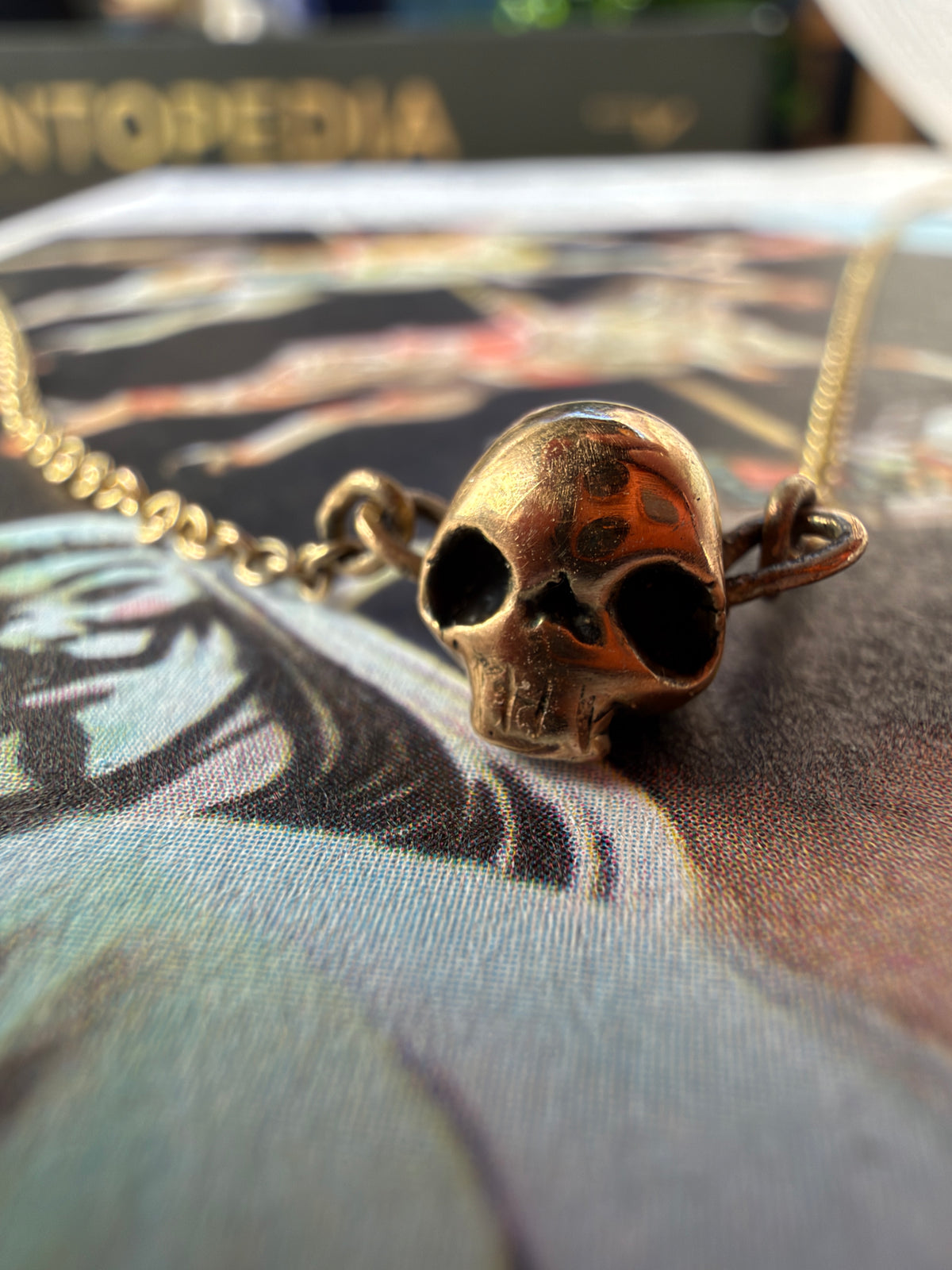 A close-up of a gold skull pendant necklace resting on a the V for vendetta graphic novel . The pendant has a warm gold finish with dark oxidized eye sockets and is attached to a matching chain.