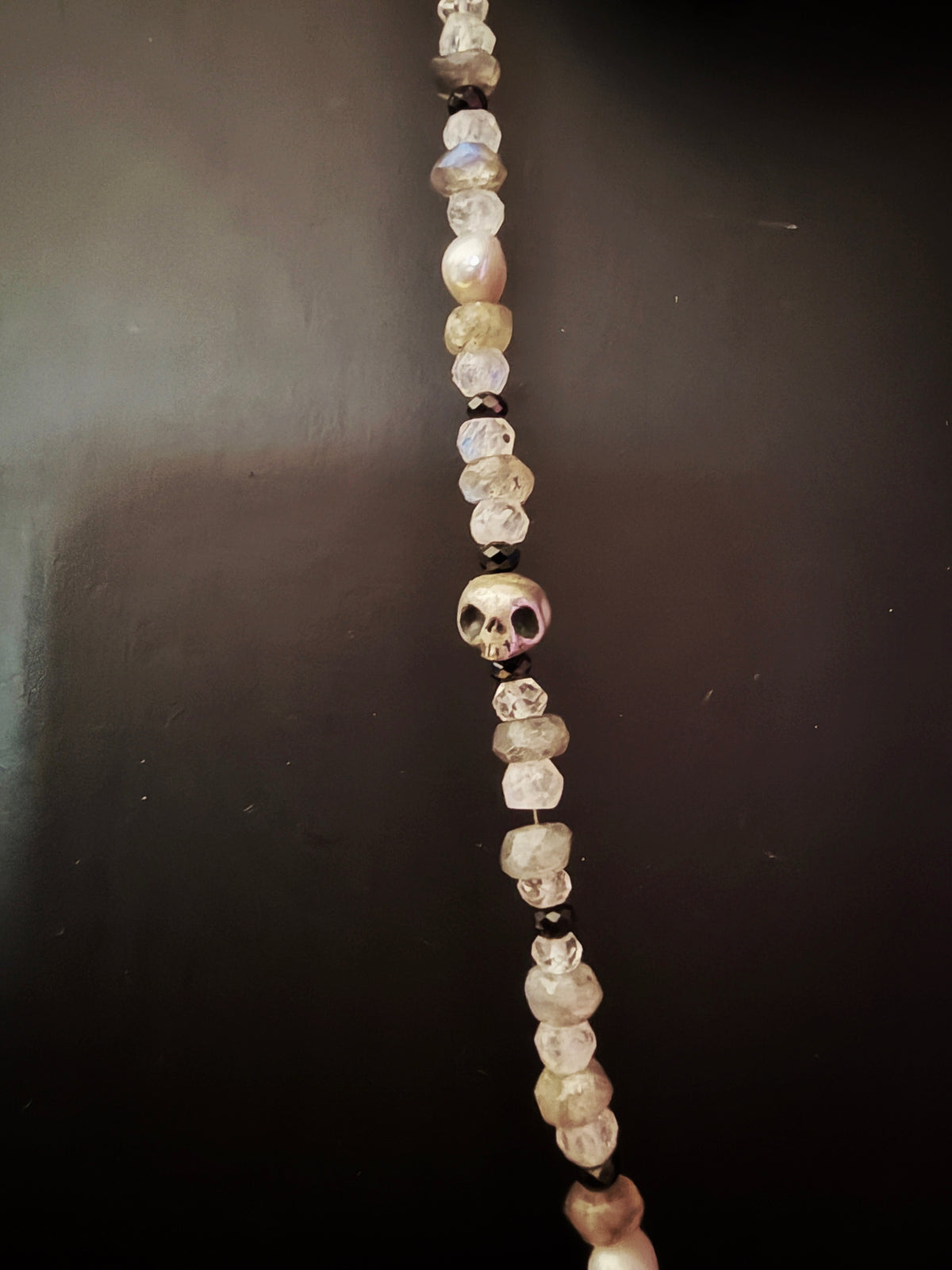 A beaded necklace featuring a central skull charm surrounded by faceted clear beads, translucent white beads, and iridescent pearl beads on a dark background.