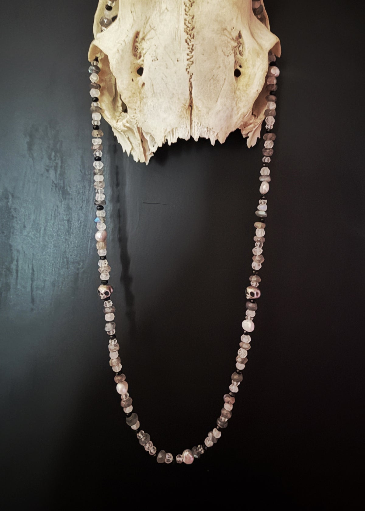 A beaded necklace made of faceted pink, white, and dark gray beads, draped below a large animal skull against a dark black background.