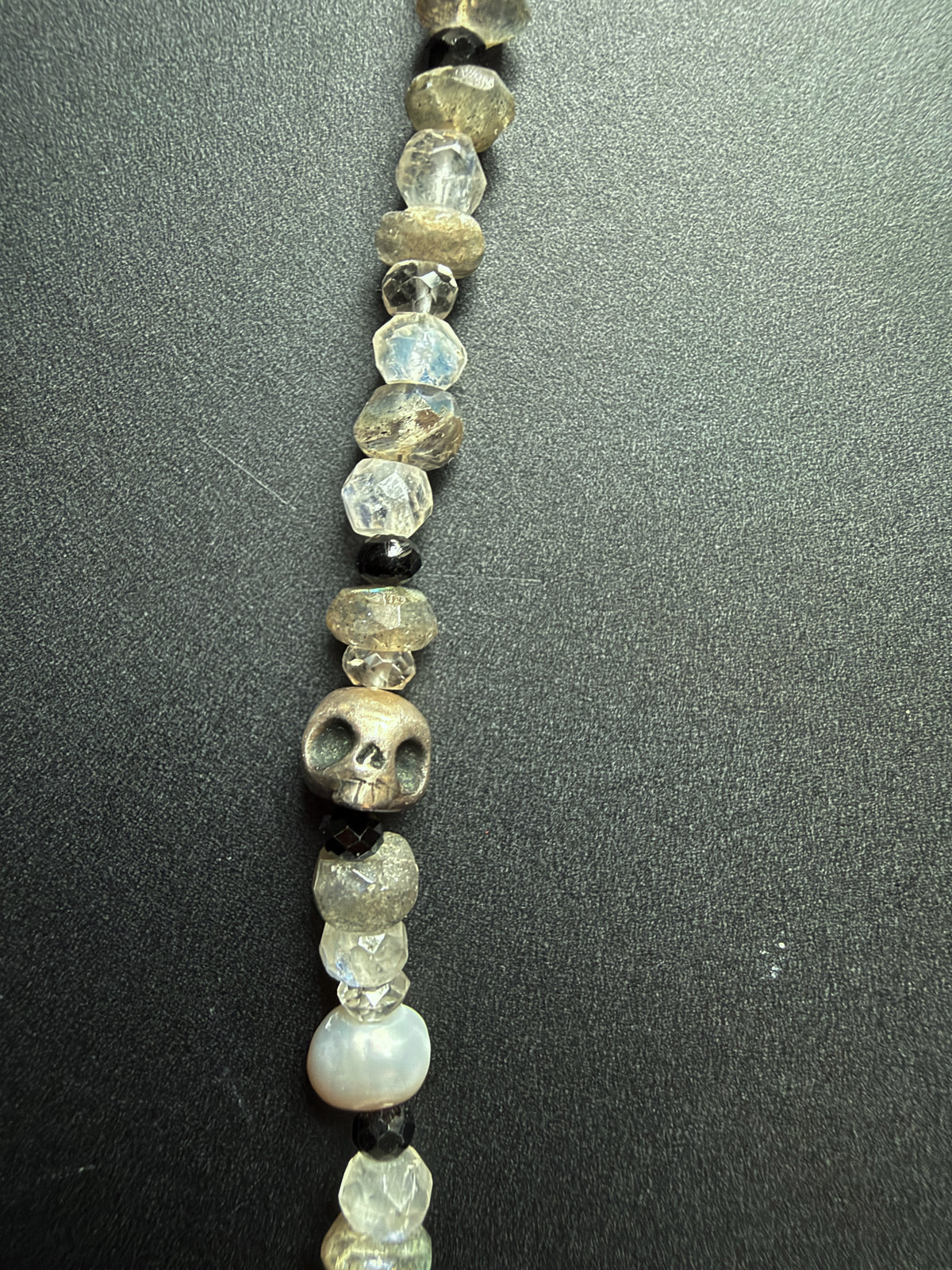 A close-up of a beaded strand featuring faceted clear quartz beads, a carved skull bead, small black beads, and a white mother-of-pearl bead, laid out on a dark gray textured background.