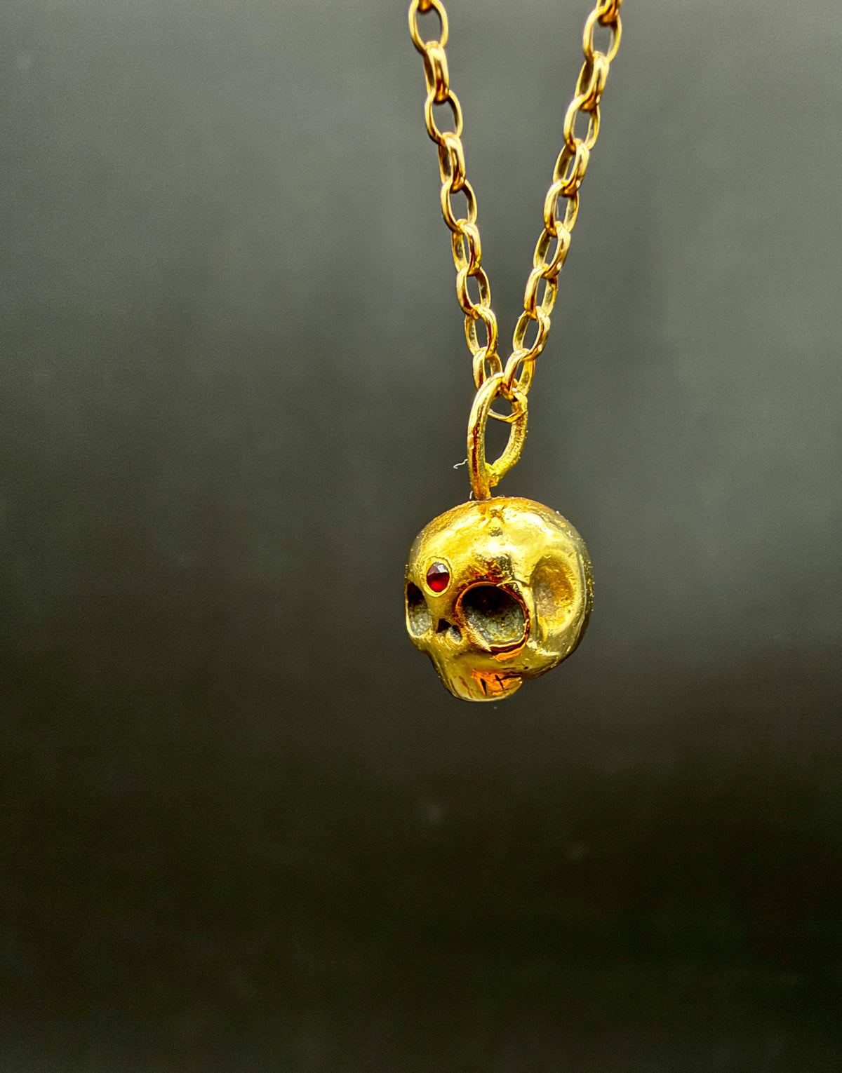 A gold skull pendant necklace with a small ruby gemstone eye, hanging from a gold link chain against a dark gray background.