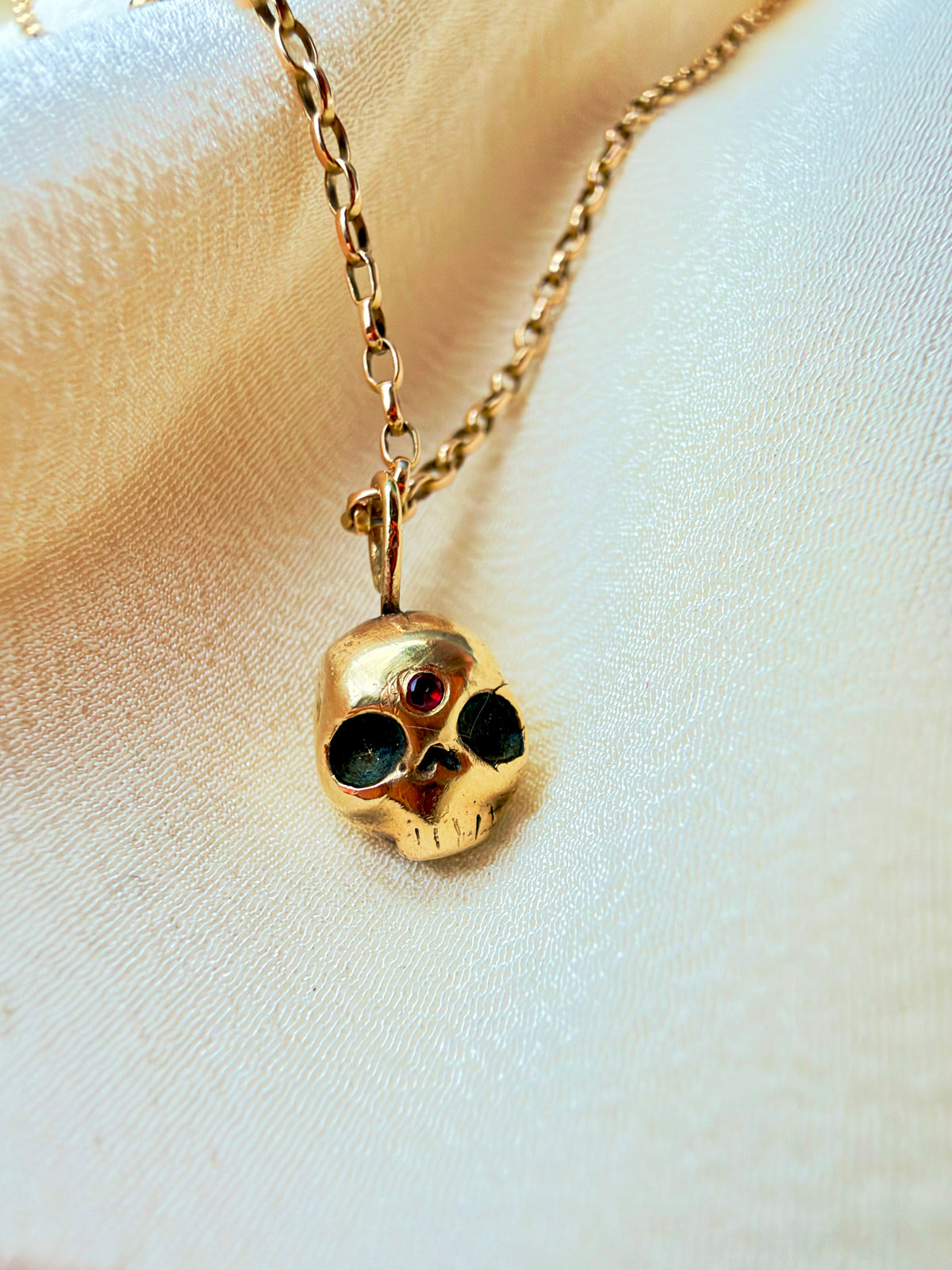 A gold skull pendant necklace with a small ruby gemstone in the center, resting on a textured white fabric background.