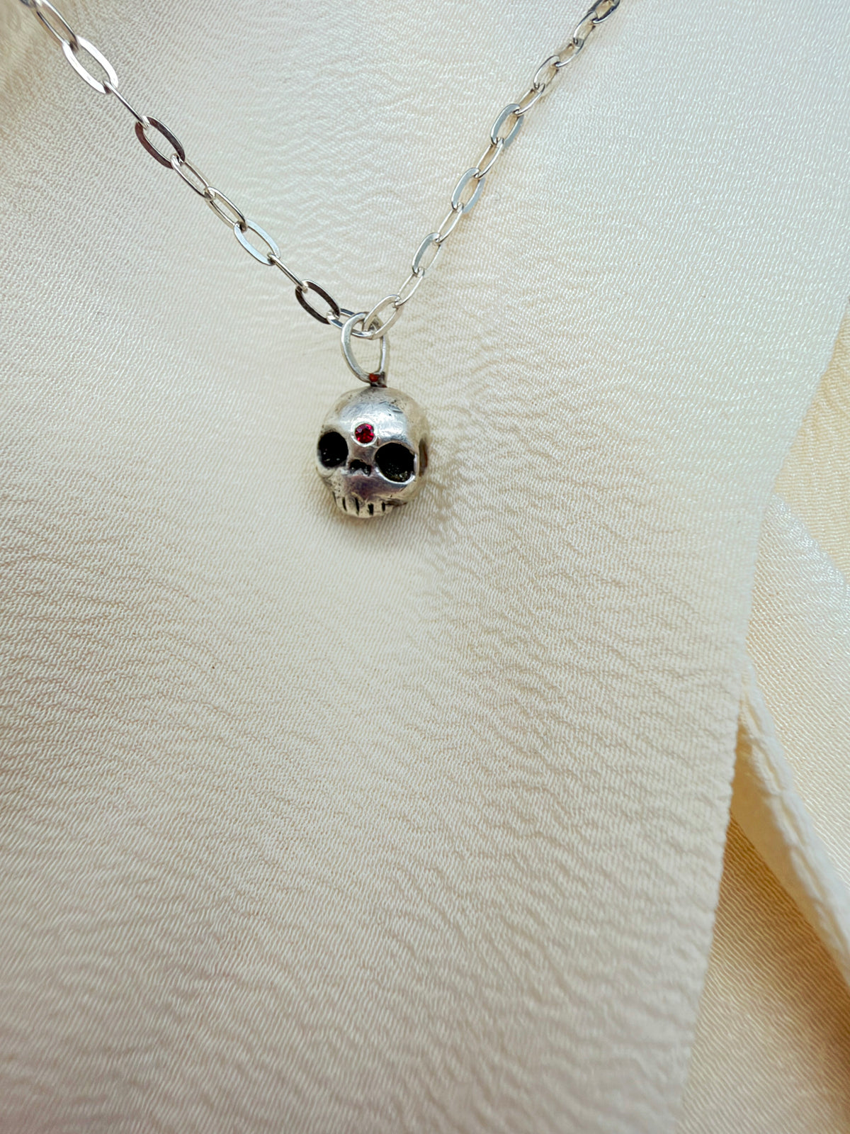 A silver skull pendant necklace with black enamel details and a small red gemstone on the forehead, displayed on a cream-colored textured fabric background.