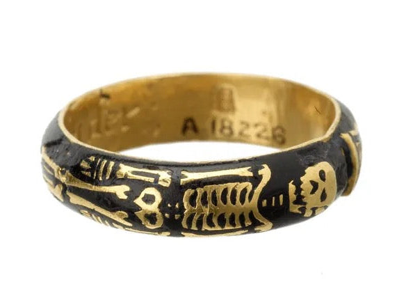 A vintage gold ring featuring a black enamel inlay with gold-gilded skeleton and skull motifs, shown on a plain white background.